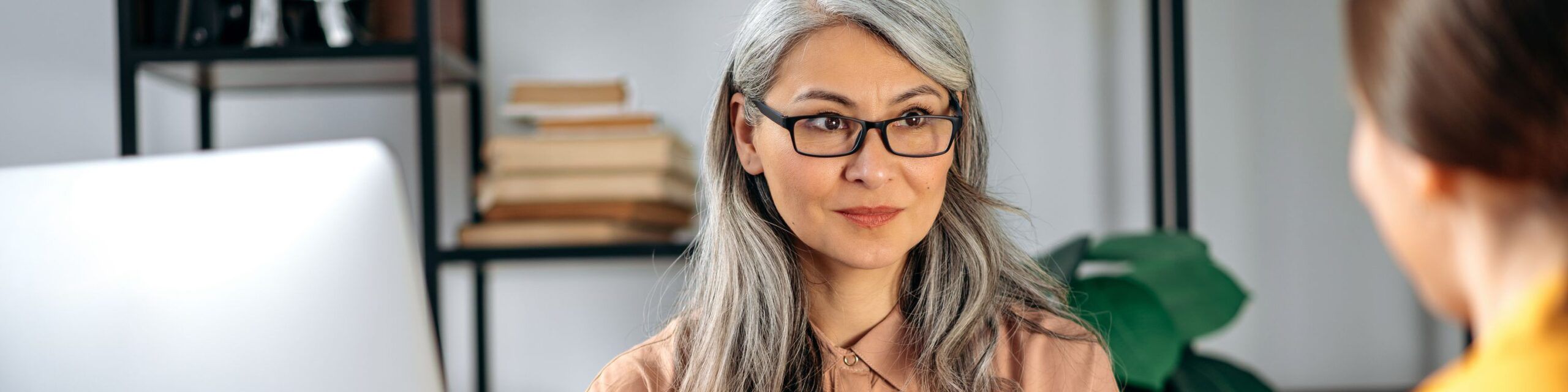 A woman with long gray hair and glasses, wearing a peach-colored shirt, sits in an office setting with bookshelves in the background, attentively listening to another person who is partially visible.