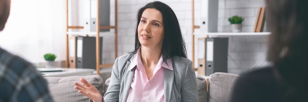 A woman with long dark hair and a light pink blouse under a grey blazer sits on a sofa, engaged in conversation with two people whose faces are not visible. Shelves with files, potted plants, and an Auto Draft sign in the background give the setting a professional atmosphere.