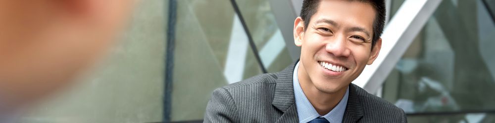 A man in a suit, smiling, is sitting outdoors near a modern glass building.