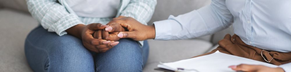 A therapist gently holds a person's hands, offering comfort and support. The person sits on a couch, partially off-frame, while the therapist holds a clipboard, suggesting a counseling session.