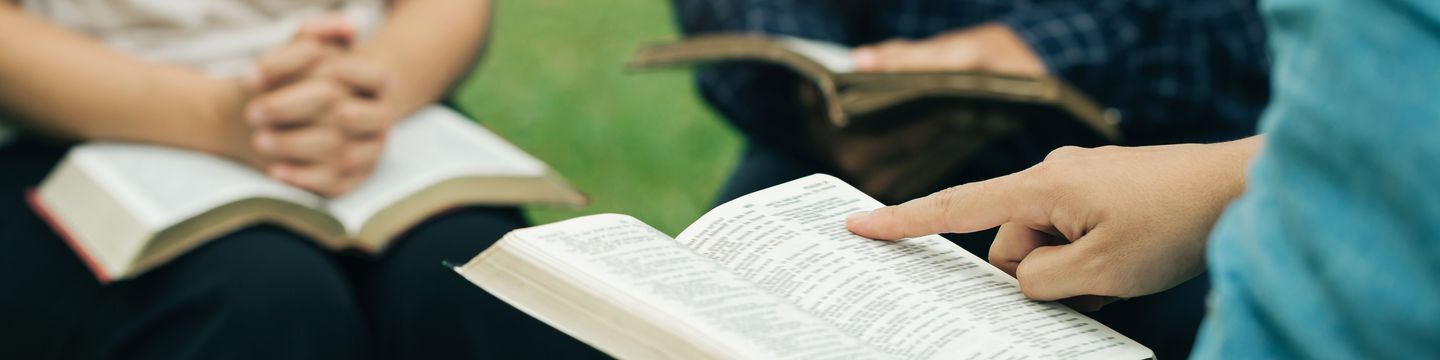 A close-up of a person reading from an open book on forensic psychology, pointing at a passage. Two others sit nearby with books open, one with hands clasped. They appear to be engaged in a group reading or discussion outdoors, delving into the nuances of criminal minds.